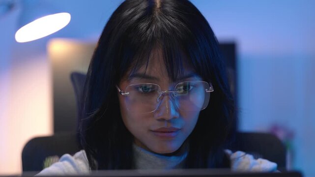 Young Asian woman developer wearing glasses sitting at a workstation late at night while typing program code on her computer with desk lamp and bright monitor behind her