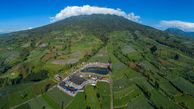 Aerial view of Embung Bansari reservoir on the slopes of Mount Sindoro surrounded by lush agricultural terraces and fields under a blue sky, Temanggung, Central Java, Indonesia.