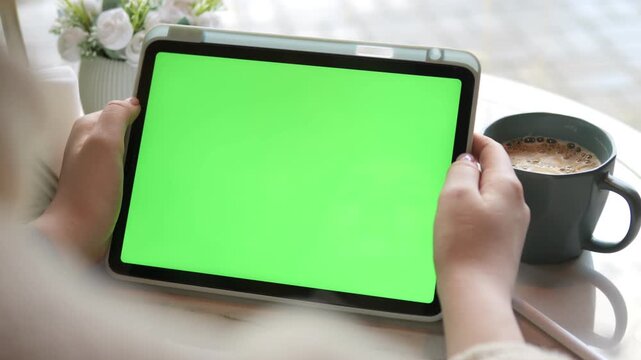 Person holds tablet with green screen. Coffee cup sits beside device. White flowers in vase nearby. Soft light fills scene