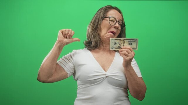 Senior woman smiling while holding a twenty dollar banknote points thumb to chest in studio; pride savings.