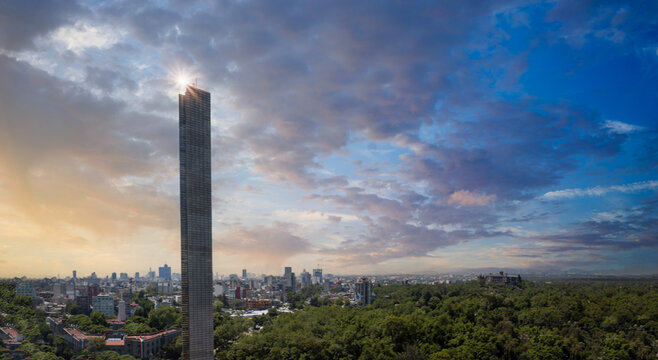 Aerial view of the slender Torre Reforma skyscraper with a sun flare overlooking the lush Chapultepec park and urban skyline under a cloudy sky in Mexico City, Mexico City, Mexico.