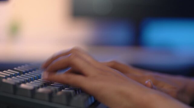 Side view close up of female Asian developer hands typing rapidly on a black computer keyboard with shallow depth of field inside a dim blue modern office environment