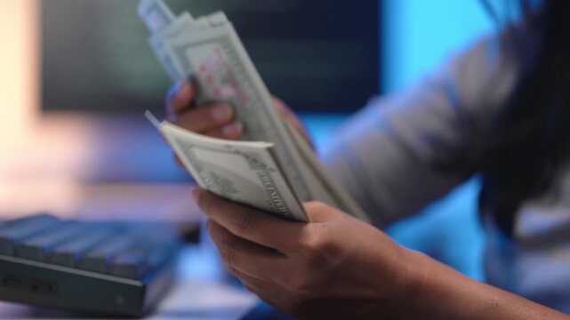 Close up of young Asian woman hands holding and counting a thick stack of US one hundred dollar bills next to a black calculator placed on her modern office desk