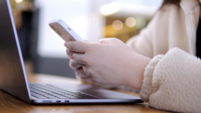Close-up of hands using smartphone beside laptop. Person wears light-colored fleece sleeves. Background softly blurred with warm bokeh lights. Wooden desk surface visible under devices