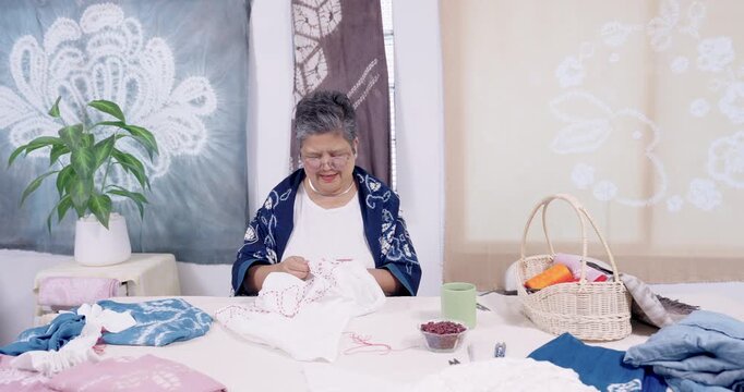 Senior Asian craftswoman continues stitching shibori pattern on cloth at home studio table, tools and dyed samples nearby, heritage craft revival video content for small business marketing now.