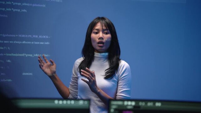 Young Asian female software engineer in a white sweater standing in front of a giant blue digital screen filled with programming code while presenting a lecture