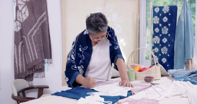 Retired Asian woman entrepreneur leans forward, marks fabric pattern lines on worktable, planning shibori resist stitching before indigo dyeing, home studio craft business process for custom order.