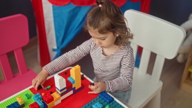 Girl stacking colorful toy blocks with hands at a kids table in a playroom building, constructing a small tower and sorting bricks; concentration creativity.
