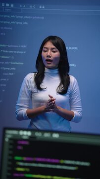 Vertical full body portrait of young Asian woman programming lecturer in a white turtleneck gesturing with both hands while explaining code inside a modern tech classroom