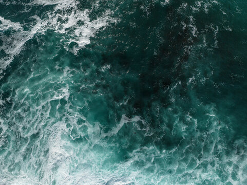 Aerial view of turquoise ocean waves crashing with white foam and dark kelp visible beneath the surface at La Jolla Cove in San Diego, California, United States.
