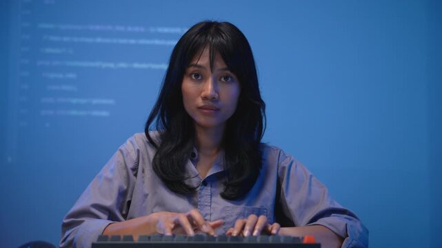 Young Asian woman software developer in a blue shirt focused on typing program code on her mechanical keyboard with bright lines of code projected on the wall behind her