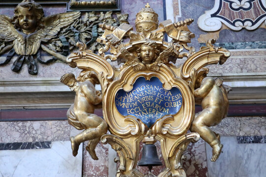 Sfarzoso stemma araldico papale in legno intagliato e dorato con putti e tiara sopra una campana bronzea all'interno della Basilica di San Giovanni in Laterano