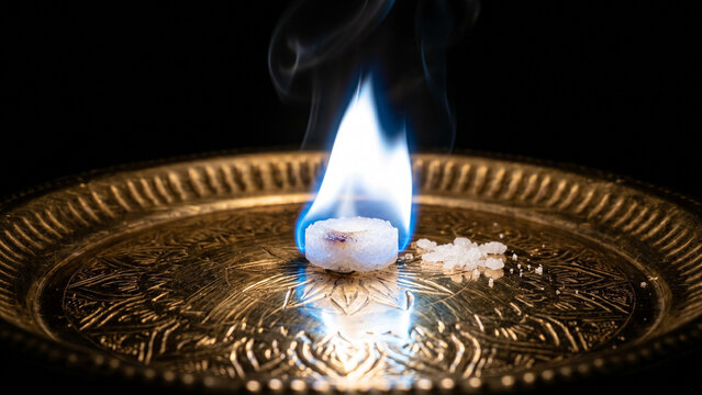 Burning camphor tablet with vivid white-blue luminous smokeless flame on polished brass puja plate with caustic reflections on black background