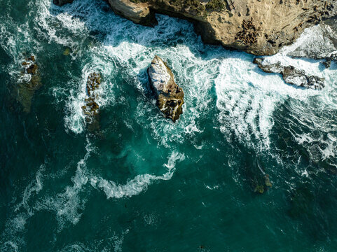 Aerial view of La Jolla Cove with turquoise ocean waves crashing against rugged rocks and sea lions resting on the shore in San Diego, California, United States.