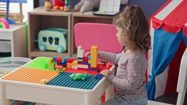 Toddler girl stacking colorful plastic building blocks and reaching with small hands at a play table in a building filled with toys and a play tent; concentration.