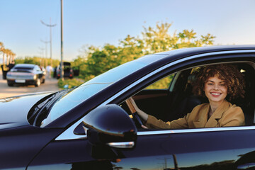 Fototapeta premium A smiling woman sits behind the wheel of a sleek car, looking out from the window with a carefree lifestyle vibe, enhanced by retro film color and warm, sunny tones.