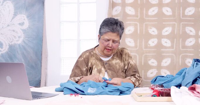 Senior Asian woman entrepreneur works beside laptop, uses seam ripper, cuts red resist stitching, unravels thread from blue cotton shirt, organizes shibori pieces for online shop order in home studio.