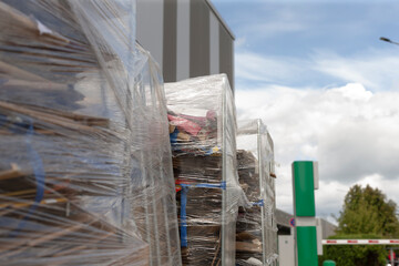 Pallets wrapped in plastic positioned along service station perimeter, green signage and trees in background, contrast between industrial packaging and roadside © Viktar