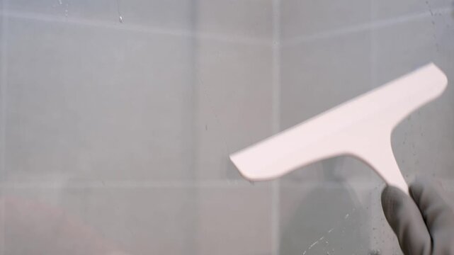 A close-up of a hand in a rubber glove cleaning the glass in shower stall with a scraper.