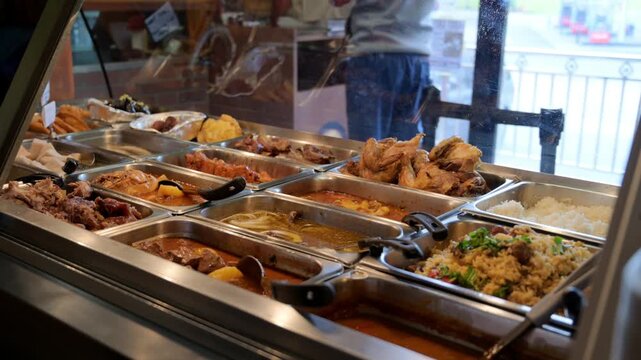 A steady close-up shot of various fried appetizers and snacks kept warm in silver trays inside a professional heated glass display at a food market.