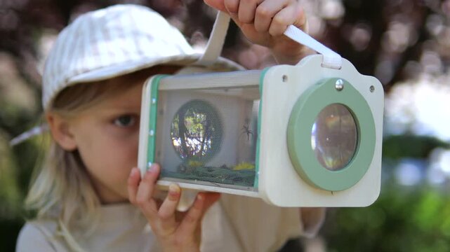 Young girl with a hat exploring the outdoors, looking intently through a magnifying bug viewer shaped like a camera, fostering curiosity and a love for nature and entomology in early childhood
