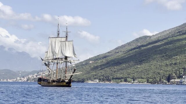 The frigate Shtandart, a replica of an 18th-century sailing ship, glides across the calm blue waters of Montenegro's Bay of Kotor on a sunny day with green mountains in the background