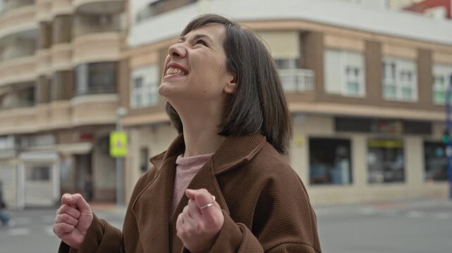 Woman in a brown coat with clenched fists, looking up and smiling on a city street by a multi story building in downtown intersection; relief hope.