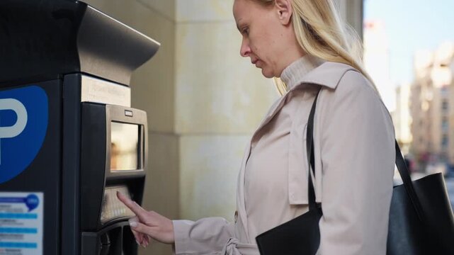 Blonde woman in beige trench coat paying for parking at a modern street payment terminal, using credit card and entering details. Urban mobility, cashless payment, and city lifestyle concept.