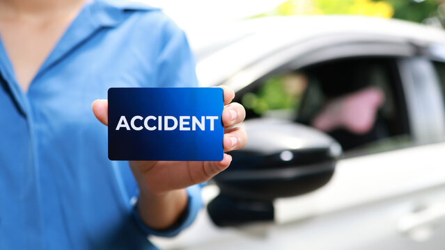 Close-up of person holding a blue card with text accident in front of a blurred car, concept of traffic collision, auto insurance claim, roadside assistance, and vehicle damage documentation.