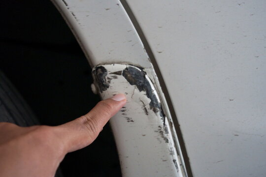 A finger pointing to a deep scratch and dent damage on a white car body bumper.