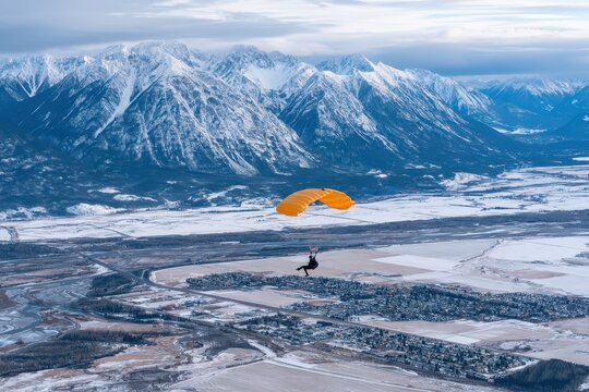 Skydiver with orange parachute descends over a snow-covered valley and majestic mountains.