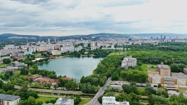 Cinematic aerial drone footage of Veľk&yacute; Draždiak lake in Petržalka, Bratislava, Slovakia. Urban cityscape, residential districts, green forest and popular recreation area in summer.