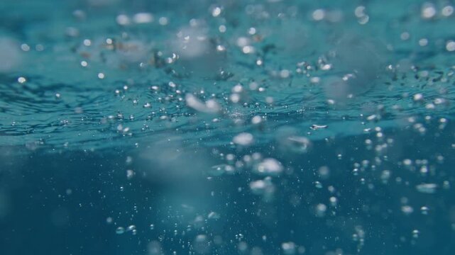 Air bubbles rise beneath ocean surface turbulence in super slow motion, revealing cavitation, water shear, and suspended microbubble movement through illuminated blue seawater.