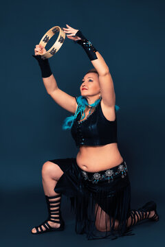 Woman stands on one knee and holds up tambourine and plays it. Mid aged adult female dancer dressed in black performance costume that was handmade - leather top and skirt, bracers and gladiator boots