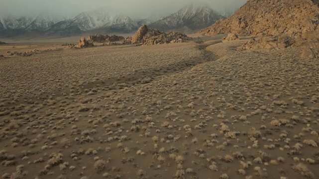 A cinematic landscape shot of the Alabama Hills in California reveals massive desert rock formations with snow-covered Sierra Nevada mountain peaks rising in the background beneath dramatic clouds. Th