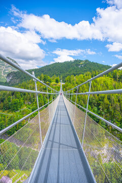 Visitors walk along the Highline 179 bridge, a suspension bridge in Austria. The bridge connects a castle ruin to a Roman fortress across a river gorge surrounded by greenery.
