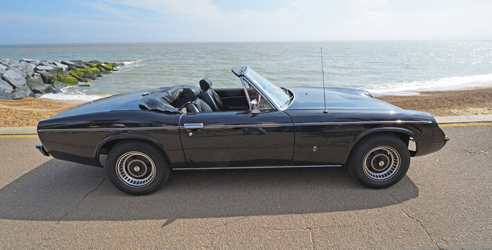 Classic black Jensen-Healey roadster parked on seafront promenade beach and sea in background. 