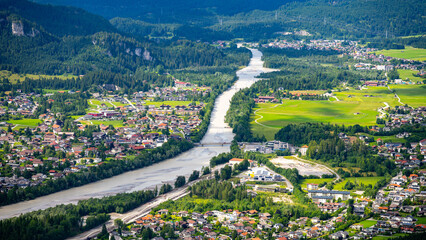 Fototapeta premium Aerial view shows the river flowing through the town of Reutte. Houses line the riverbanks, and green fields spread across the landscape. Trees and mountains surround the area.