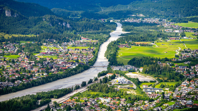 Aerial view shows the river flowing through the town of Reutte. Houses line the riverbanks, and green fields spread across the landscape. Trees and mountains surround the area.