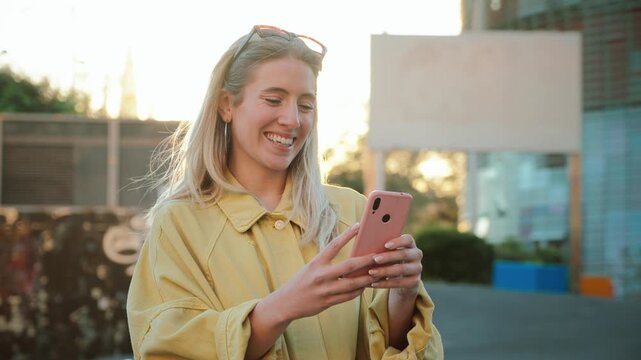 Cheerful young woman smiling as she interacts with her smartphone, showcasing modern technology and social connection in a delightful manner under bright sunlight.