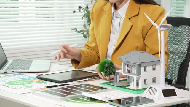 Businesswoman reviewing renewable energy and green building plans at desk with house model and wind turbine, sustainability concept, modern office, selective focus.