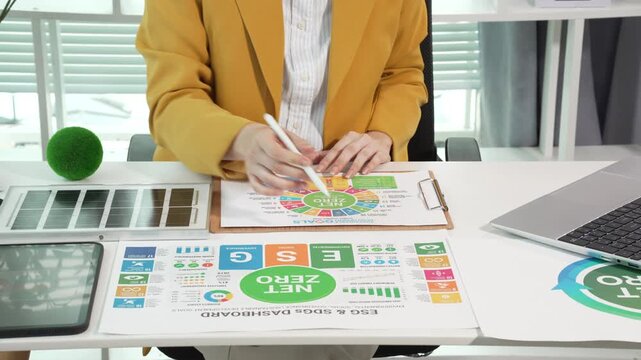 Business professional at desk reviewing sustainability report with net zero and sdg icons, using laptop and clipboard in bright modern office.