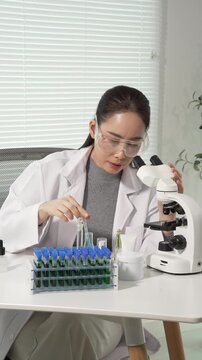 Scientist in lab coat and safety goggles using microscope at lab bench with test tubes and pipette, conducting research in bright modern laboratory.