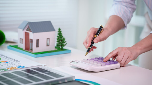 Person calculating home mortgage costs at desk with calculator and model house, financial documents and solar panel sample, real estate budgeting and sustainable housing concept.