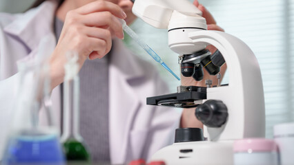 Fototapeta premium Scientist in lab coat uses pipette to place blue liquid on microscope slide beside flasks of colorful reagents in modern laboratory, close-up, shallow depth of field.