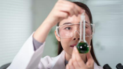 Female scientist in lab coat and safety goggles holds test tube with green liquid, focusing on experiment in bright modern laboratory, shallow depth of field. © Phushutter
