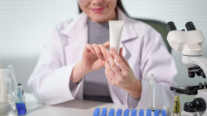 Female scientist or dermatologist in lab coat holding cosmetic cream tube in laboratory with microscope and glassware, skincare research and product testing concept. © Phushutter