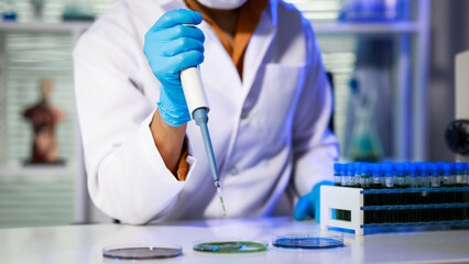 Scientist in lab coat and mask using micro pipette at bench with petri dishes and test tubes, blue gloves, modern laboratory setting. © Phushutter