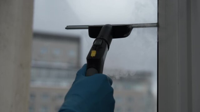 Close-up of professional cleaner using handheld steam cleaner with squeegee attachment to wash dirty glass window, providing deep sanitation and maintenance for modern apartment building, slow motion.