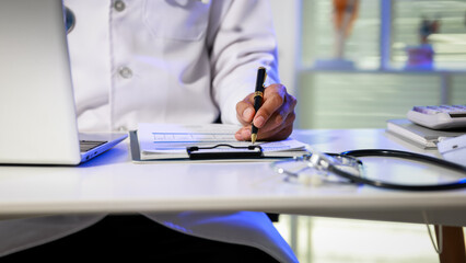 Fototapeta premium Doctor in white coat writing on clipboard at desk with laptop and stethoscope in foreground, clinical office setting, healthcare documentation and diagnosis concept.